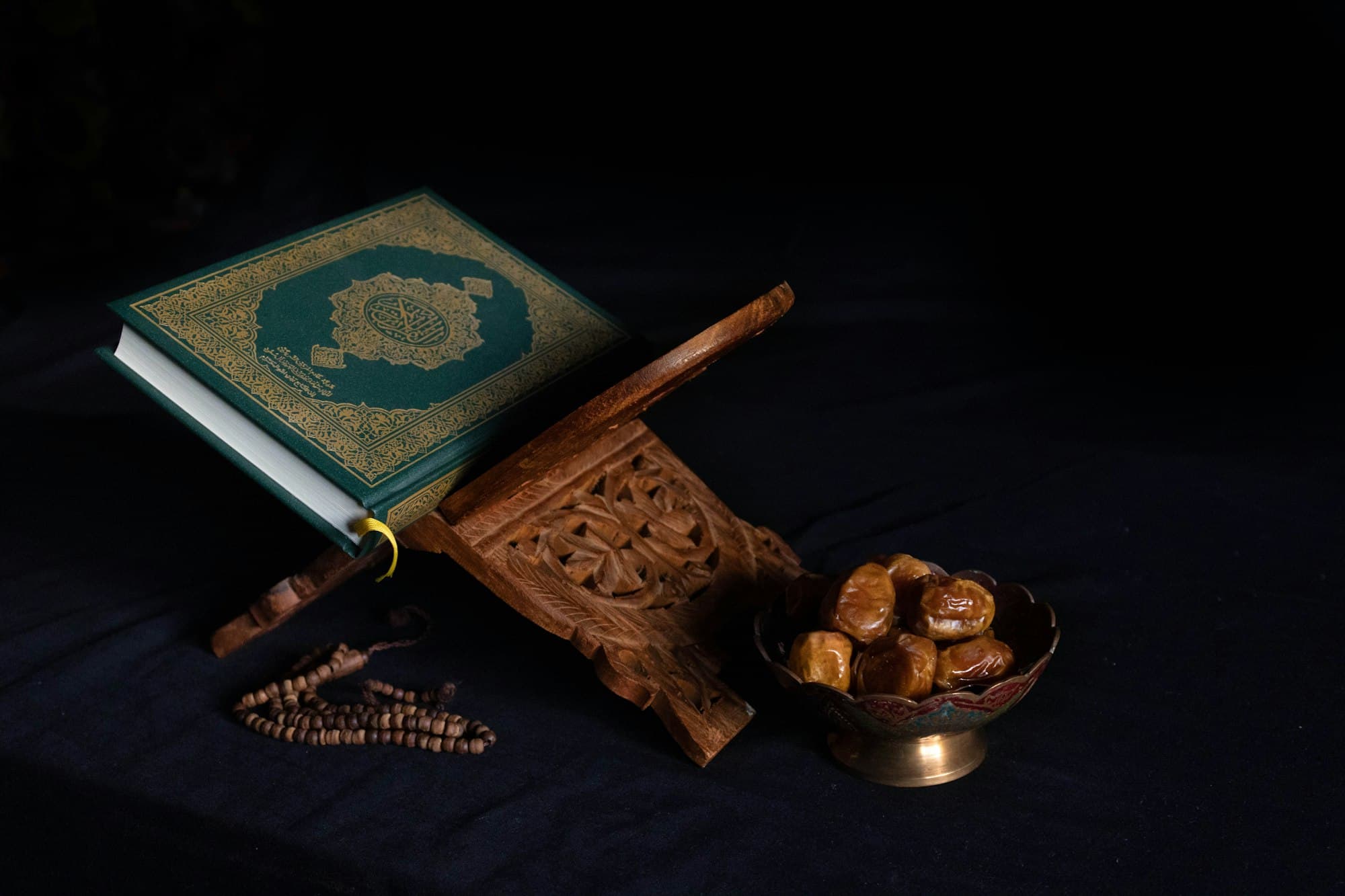 A green Quran on a carved wooden stand beside dates and prayer beads